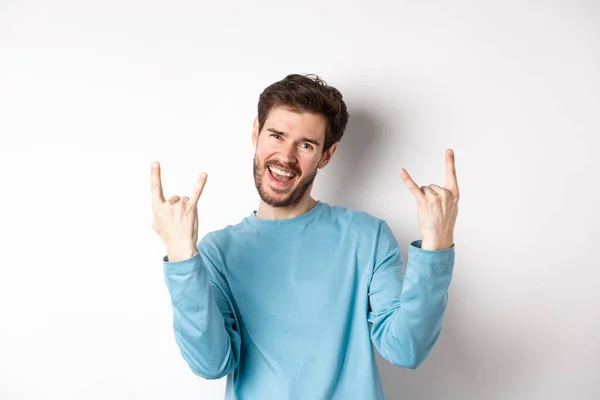 Emocionado hombre guapo celebrando, divirtiéndose y mostrando la roca en los cuernos gesto, disfrutando de la fiesta, sonriendo a la cámara, de pie sobre fondo blanco —  Fotos de Stock