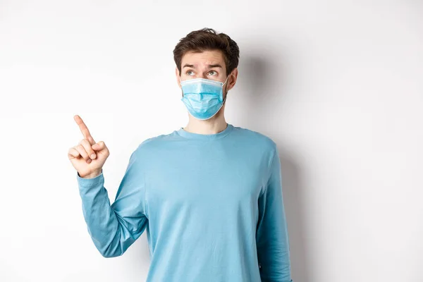 Coronavirus, hälsa och karantän koncept. Nyfiken kille i medicinsk mask, peka och titta på övre vänstra hörnet banner, står på vit studio bakgrund — Stockfoto