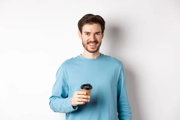 Hombre alegre bebiendo café de la cafetería para llevar y sonriendo, de pie con taza de papel en fondo blanco — Foto de Stock