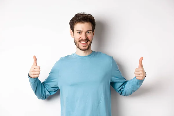 Jovem alegre mostrando os polegares para cima e sorrindo, recomendo um bom produto, elogiando a escolha, de pé sobre fundo branco — Fotografia de Stock