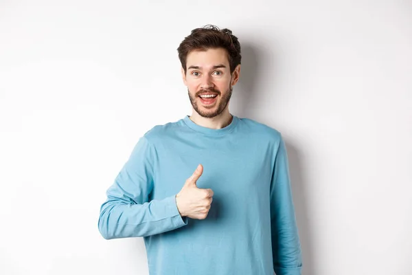 Jovem alegre concorda e diz sim, mostra o polegar na aprovação, sorrindo feliz para a câmera, de pé sobre o fundo branco — Fotografia de Stock