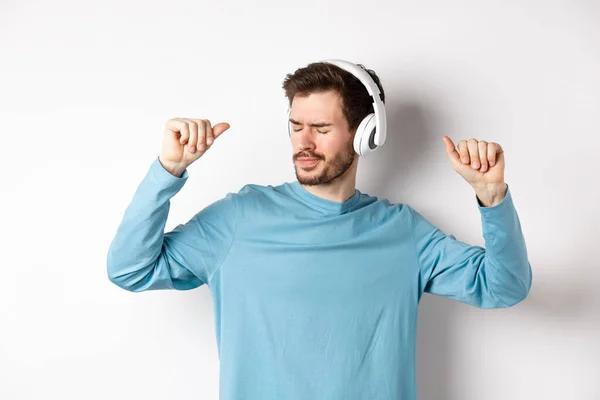 Happy young man having fun in headphones, dancing while listening music in wireless earphones, white background — Stock Photo, Image