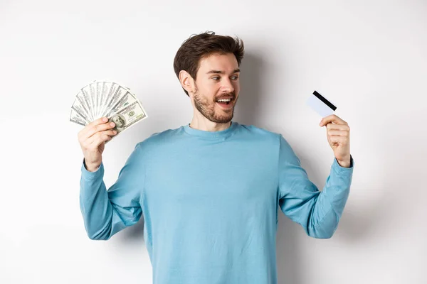 Hombre barbudo guapo elegir entre el dinero y la tarjeta de crédito de plástico, el pago con efectivo o sin contacto, de pie sobre fondo blanco — Foto de Stock