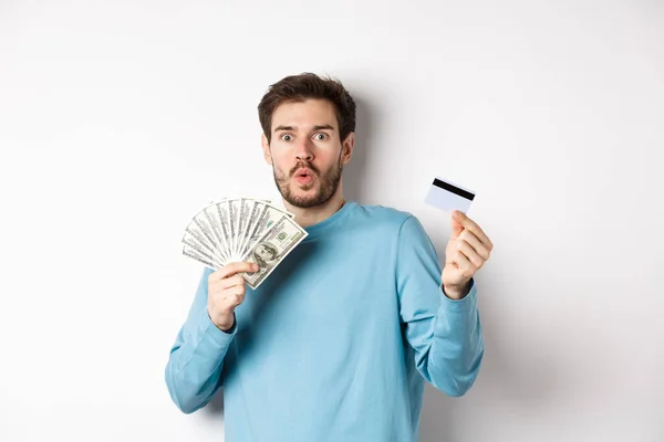 Sorprendido joven mostrando dinero en dólares y tarjeta de crédito de plástico, jadeando y diciendo wow con la cara sorprendida, de pie sobre fondo blanco — Foto de Stock