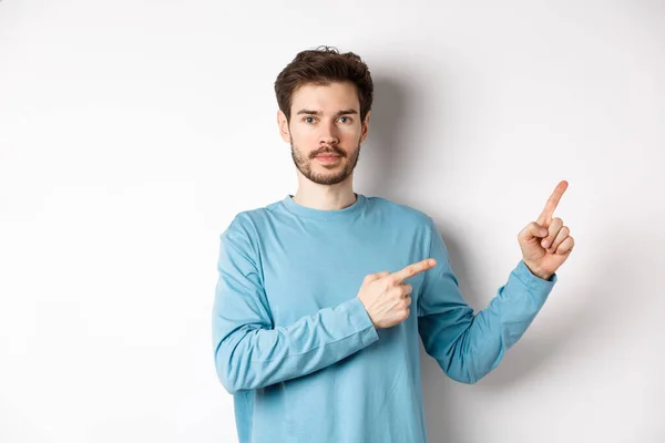 Portrait d'un homme caucasien sérieux avec la barbe pointant droit sur le logo et regardant la caméra, debout dans des vêtements décontractés sur fond blanc — Photo