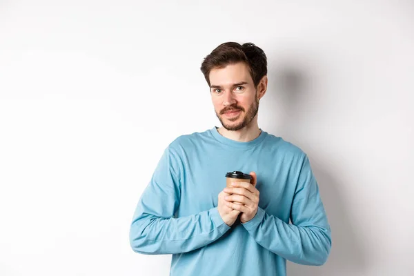 Snygg kaukasisk man uppvärmning händer med kopp kaffe, ler mot kameran och ser ömt, står på vit bakgrund — Stockfoto