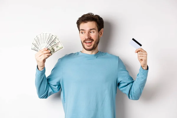 Emocionado hombre barbudo elegir entre el dinero y la tarjeta de crédito de plástico, el pago con dinero en efectivo o sin contacto, de pie sobre fondo blanco — Foto de Stock