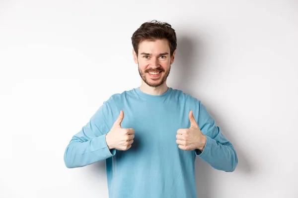 Sorrindo homem bonito mostrando polegares para cima, como e aprovar boa escolha, elogiando ou recomendando algo, de pé sobre fundo branco — Fotografia de Stock