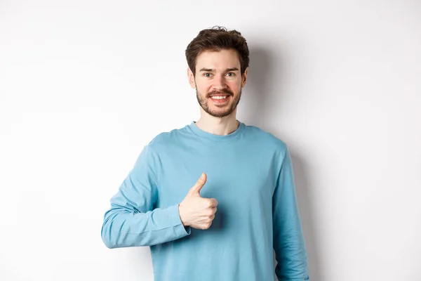 Hymyilevä miesmalli rento paita osoittaa peukalo ylös, hyväksyä ja suositella tuote, kehua erinomainen valinta, seisoo valkoisella taustalla — kuvapankkivalokuva
