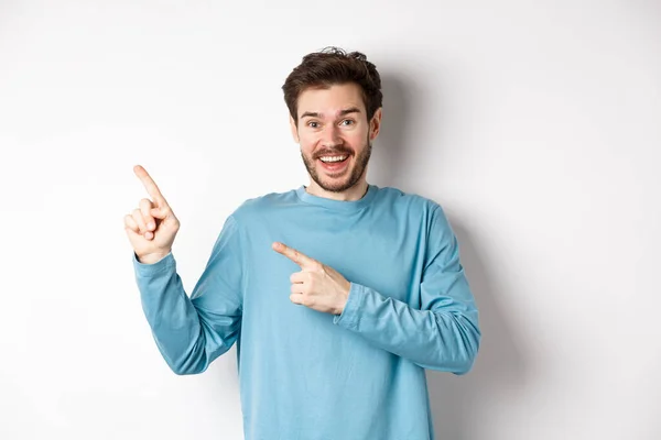Happy caucasian man in casual blue sweatshirt, pointing fingers at upper left corner, showing link or logo on white background, smiling at camera — Stock Photo, Image