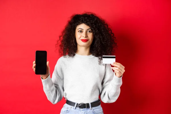 Mujer moderna joven con el pelo rizado que muestra la tarjeta de crédito de plástico y la pantalla del teléfono móvil, demostrando aplicación de compras en línea, de pie sobre fondo rojo — Foto de Stock