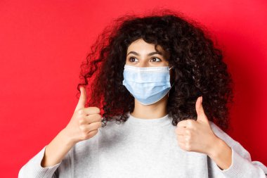 Covid-19 ve pandemik konsept. Kıvırcık saçlı, tıbbi maske takan, başparmağını kaldırıp boş alana bakan, kırmızı arka planda kalan beyaz bir kadının yakın çekimi.