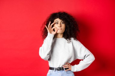 Image of cute girl with curly hair making promise to stay quiet, zipping lips, making seal, hiding a secret, standing dumb on red background