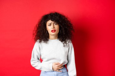 Kadın hasta, eğiliyor ve karnına dokunuyor, karın ağrısı ya da regl sancısı çekiyor, kırmızı arka planda duruyor.