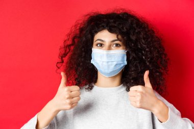 Sağlıklı insanlar ve covid-19 konsepti. Kıvırcık saçlı, salgın sırasında kendini korumak için tıbbi maske takan gülümseyen kadının yakın plan portresi.