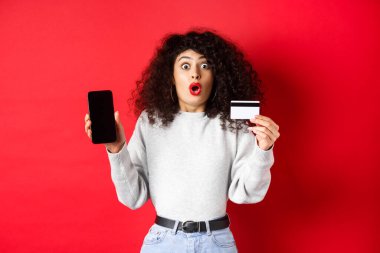 Kredi kartı ile heyecanlı bir kadın boş bir akıllı telefon ekranı gösteriyor, hayret içinde, kırmızı arka planda duruyor.