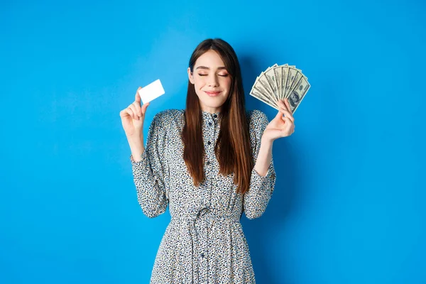 Compras. Mujer sonriente feliz mirando satisfecho con los ojos cerrados, mostrando billetes de dólar de dinero y tarjeta de crédito de plástico, de pie de ensueño sobre fondo azul — Foto de Stock