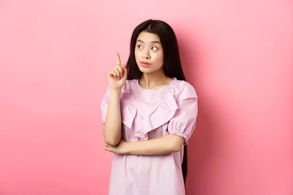 Όμορφη Ασιάτισσα κοπέλα που σηκώνει το δάχτυλο και κοιτάζει αριστερά στο λογότυπο, έχοντας μια ιδέα, βρήκε λύση, στέκεται απέναντι σε ροζ φόντο — Φωτογραφία Αρχείου