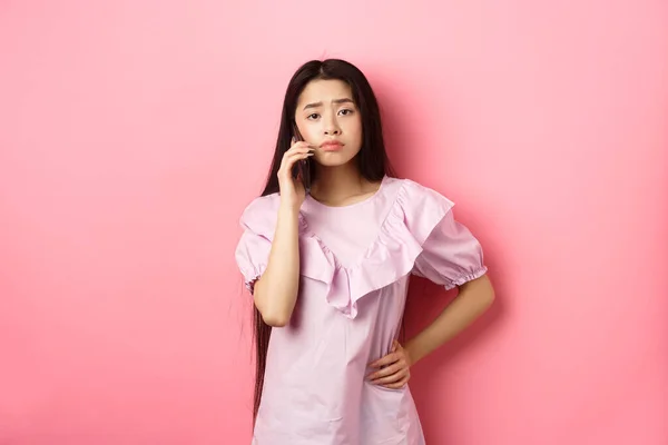 Szomorú ázsiai lány hív valakit, tartja a telefont, és beszél, áll ideges ellen rózsaszín háttér — Stock Fotó