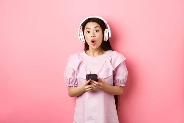 Femme asiatique excitée dans les écouteurs sans fil dire wow, tenant téléphone portable et regardant amusé à la caméra, debout sur fond rose — Photo
