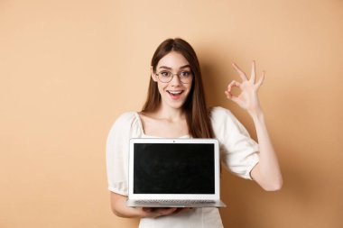 E-ticaret. Gözlüklü heyecanlı genç bir kadın iyi bir tabela ve dizüstü bilgisayar gösteriyor. İnternet promosyonu öneriyor, bej arka planda duruyor.