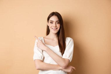 Romantik elbiseli genç bir bayan model sol üst köşeyi işaret ediyor, kameraya gülümsüyor, seçimini gösteriyor, ürün öneriyor, bej arka plan