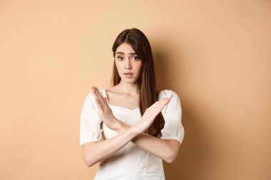 Kız hayır diyor. Şüpheci genç kadın aynı fikirde değil ya da eylemi yasaklıyor, çapraz duruş hareketi gösteriyor, işaret vermiyor, kötü bir fikri onaylamıyor, bej arka planda duruyor.
