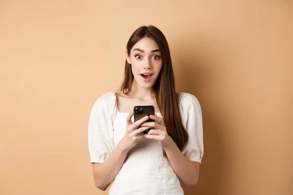 Отличные новости по телефону. Удивительная молодая женщина сказать ничего себе после прочтения онлайн объявление на смартфоне, стоя на бежевом фоне — стоковое фото