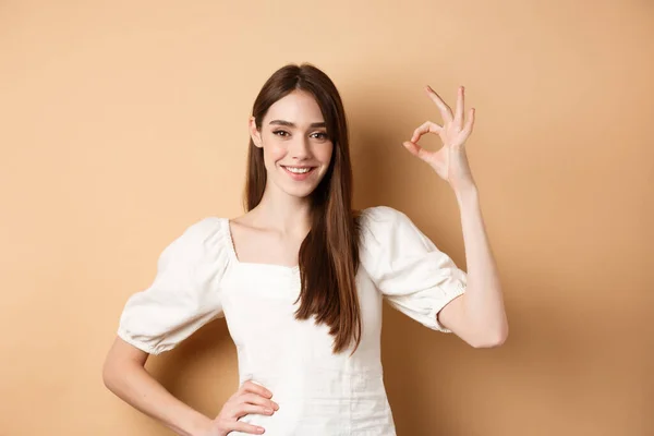 Πολύ ωραία. Χαμογελώντας χαριτωμένο γυναίκα σε λευκή μπλούζα δείχνει εντάξει σημάδι στην έγκριση, όπως και επαίνους εξαιρετική επιλογή, στέκεται ικανοποιημένος σε μπεζ φόντο — Φωτογραφία Αρχείου