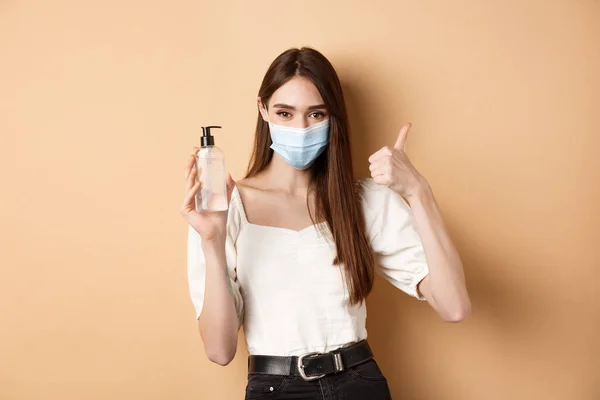 Covid-19 and preventive measures concept. Smiling girl in medical mask show thumb up and hand sanitizer, recommend product for disinfection, beige background — Stock Photo, Image