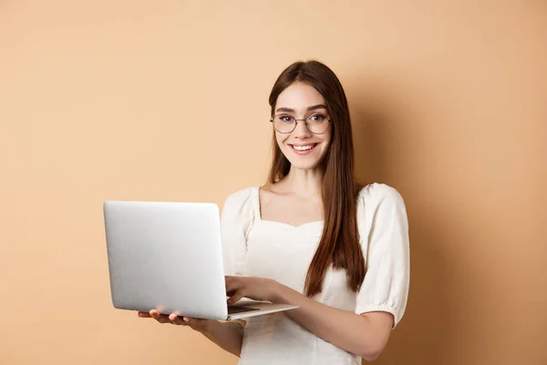 Étudiante souriante travaillant sur un ordinateur portable, portant des lunettes et regardant heureux, utilisant l'ordinateur tout en se tenant debout sur fond beige — Photo