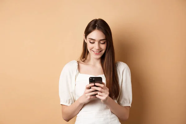 Młoda kobieta SMS-uje na telefonie komórkowym, uśmiecha się i czyta ekran smartfona, stojąc na beżowym tle — Zdjęcie stockowe