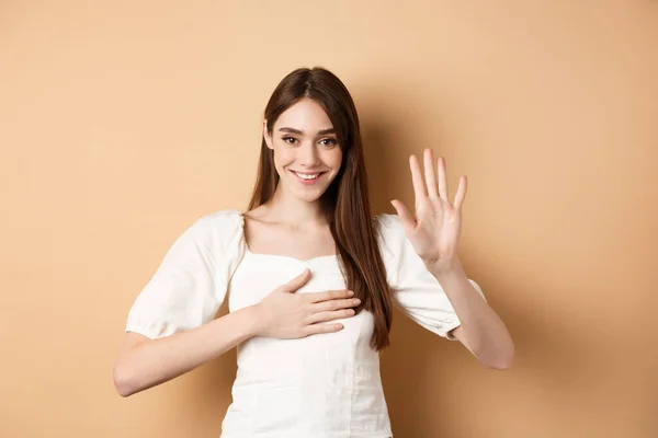 Menina sorridente bonito fazer promessa, colocar a mão no coração e dizer a verdade, sendo honesto, jurando para você, de pé sobre fundo bege — Fotografia de Stock