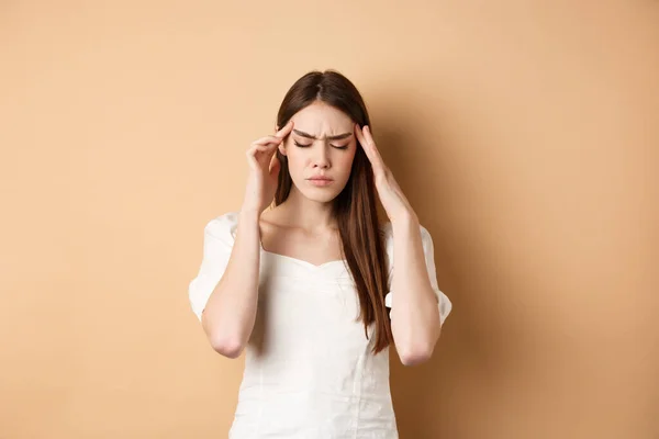 Mujer joven con dolor de cabeza, tocando las sienes de la cabeza con los ojos cerrados y la cara tensa, de pie con migraña dolorosa sobre fondo beige —  Fotos de Stock