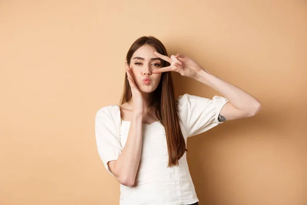 Nette Frau mit V-Zeichen und Puckerlippen zum Küssen, berührendes Gesicht mit albernem Gesichtsausdruck, stehend auf beigem Hintergrund — Stockfoto