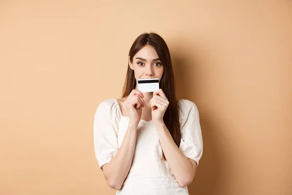 Chica excitada sosteniendo tarjeta de crédito de plástico en los labios y sonriendo, pensando en ir de compras, ir a comprar algo, de pie sobre fondo beige —  Fotos de Stock