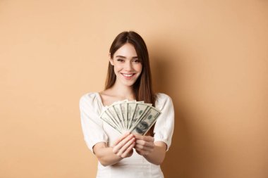 Dolar banknotları gösteren, gülümseyen, bej arka planda parası olan genç bir kadın. Kredi ve sigorta kavramı