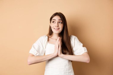 Umut dolu genç kız Tanrı 'ya yalvarıyor ve sol üst köşede rüya gibi görünüyor, dua ediyor ya da dilek tutuyor, bej arka planda duruyor.