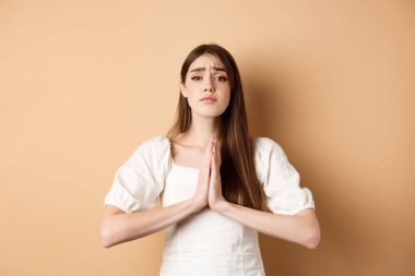 Lütfen yardım edin. Umutsuz genç bir kadın sana yalvarıyor, el ele tutuşup kameraya bakıyor, iyilik istiyor, bej bir geçmişi var.