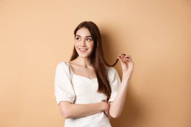 Romantik düşünceli kız, soldaki boşluğa bakıyor ve saç teli ile oynuyor, gülümsüyor, bej arka planda beyaz elbiseyle duruyor.