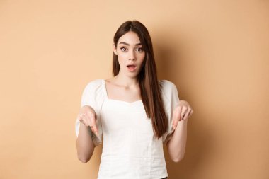 Vay canına, şuraya bak. Beyaz elbiseli hayrete düşmüş genç bir kadın, parmaklarını aşağı işaret ediyor, ağzı açık duruyor ve gözleri bej arka planda patlıyor.