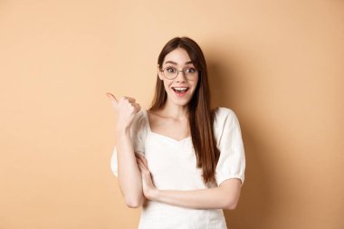 Gözlüklü heyecanlı kız logoyu işaret ediyor, şaşırmış bir şekilde gülümsüyor, tavsiye ediyor, bej arka planda duruyor.