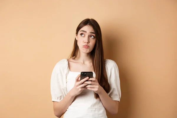 Запутавшаяся молодая женщина думает после прочтения новостей на мобильном телефоне, глядя на верхний левый угол колеблется, стоя на бежевом фоне — стоковое фото