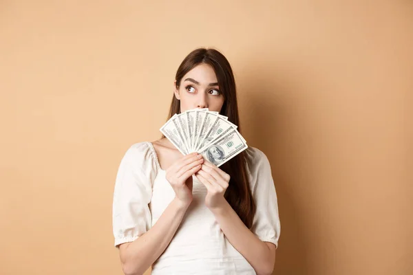 Menina cuidadosa segurando notas de dólar e olhando para o espaço vazio, pensando no que comprar em dinheiro, plano de compras, de pé sobre fundo bege — Fotografia de Stock