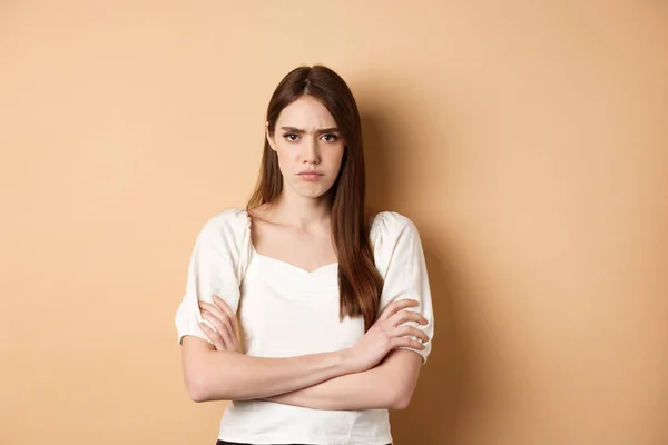 Petite amie en colère croise les bras sur la poitrine et fronçant les sourcils, regardant offensé ou déçu, vous boudant, debout fou sur fond beige — Photo