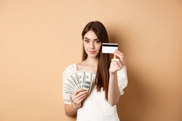 Jeune femme montrant la carte de crédit en plastique, préfèrent le paiement sans contact au lieu de billets d'un dollar, debout sur fond beige — Photo