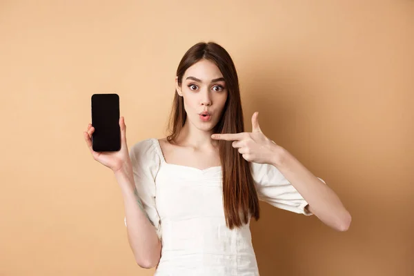 Vzrušená žena zobrazující zprávy na obrazovce, ukazující na prázdný telefon a vypadající překvapeně, stojící na béžovém pozadí — Stock fotografie