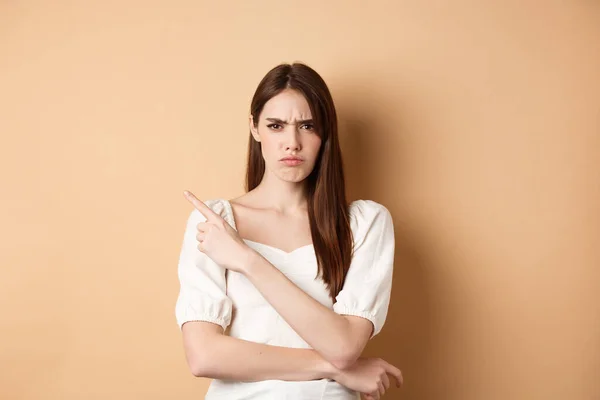 Boos fronsend meisje wijst naar lege ruimte, kijkt teleurgesteld en van streek, schold je uit of beschuldigt je, staat op beige achtergrond — Stockfoto