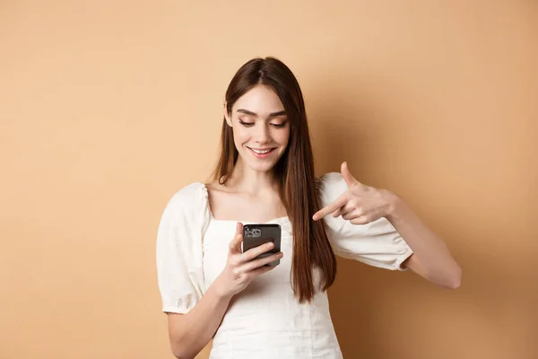 Usmívající se roztomilá dívka čtení obrazovky a ukazuje na mobilní telefon, stojící na béžové pozadí — Stock fotografie