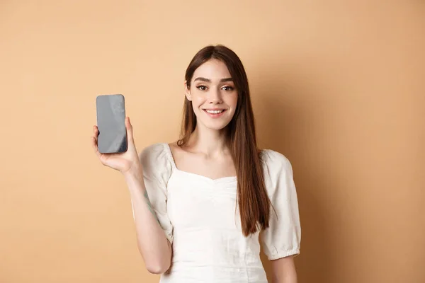 Ragazza felice mostrando schermo mobile vuoto e sorridente, dimostrare app telefono, in piedi su sfondo beige — Foto Stock